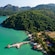 A densely forested tropical island surrounded by water, with a jetty extending into the sea and traditional village buildings along the shoreline